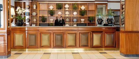 Zona de recepción de hotel con un mostrador de madera, un hombre de pie detrás y una pared decorada con jarrones y plantas.