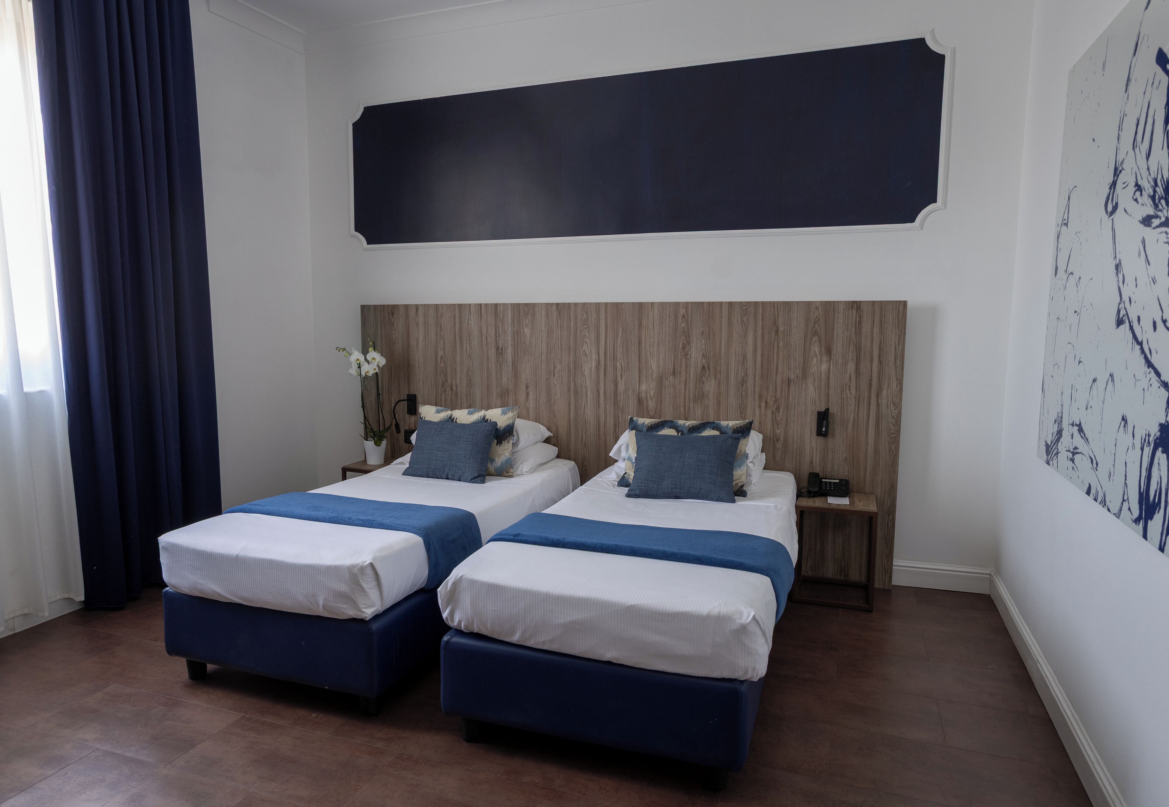 A hotel room with two beds, a wooden headboard, a nightstand, and a large abstract painting on the wall.