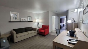 A hotel room with a sofa, a red armchair, a desk with a telephone, and a lamp. There is a door leading to a bedroom with a bed. - Country Inn & Suites by Radisson, McDonough, GA (McDonough)