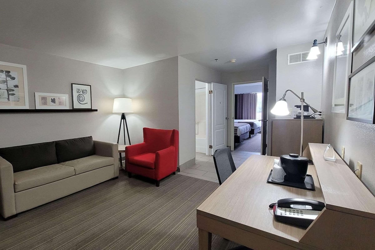 A hotel room with a sofa, a red armchair, a desk with a telephone, and a lamp. There is a door leading to a bedroom with a bed.