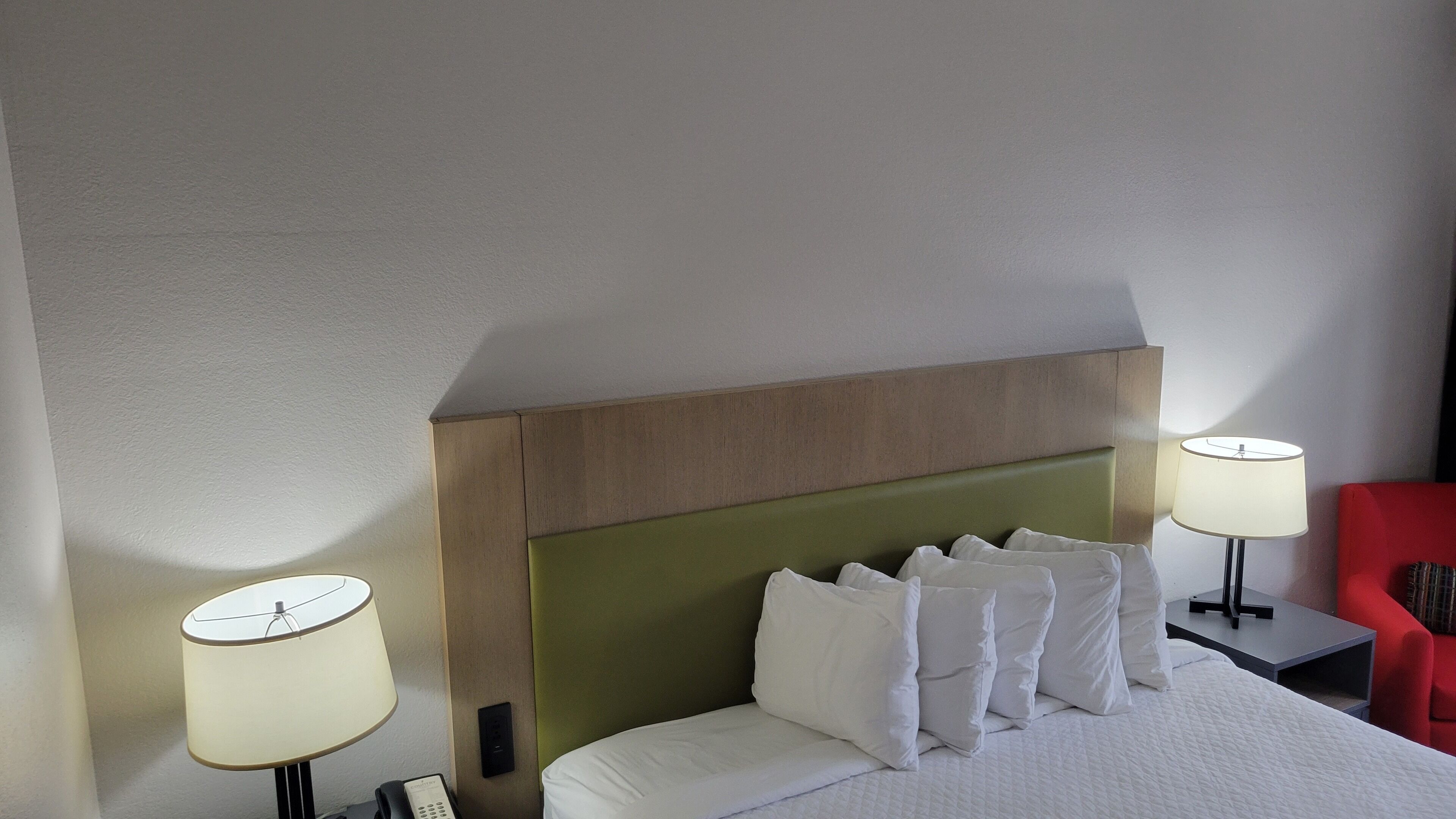 A hotel room with a bed, two bedside lamps, a red chair, and a wooden headboard.