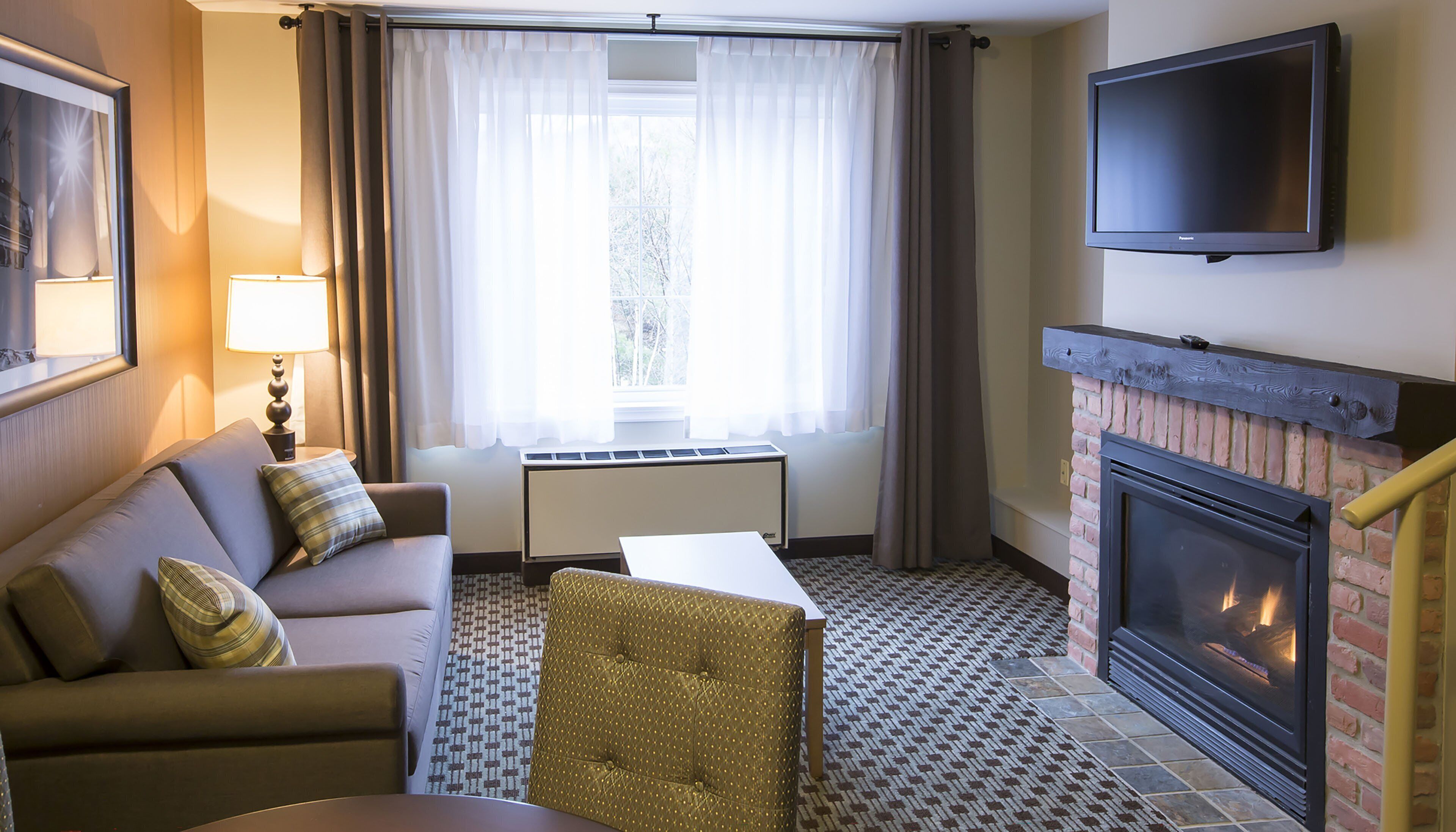 Een hotelkamer met een bank, een klein tafeltje, een open haard, een televisie en een raam met gordijnen.