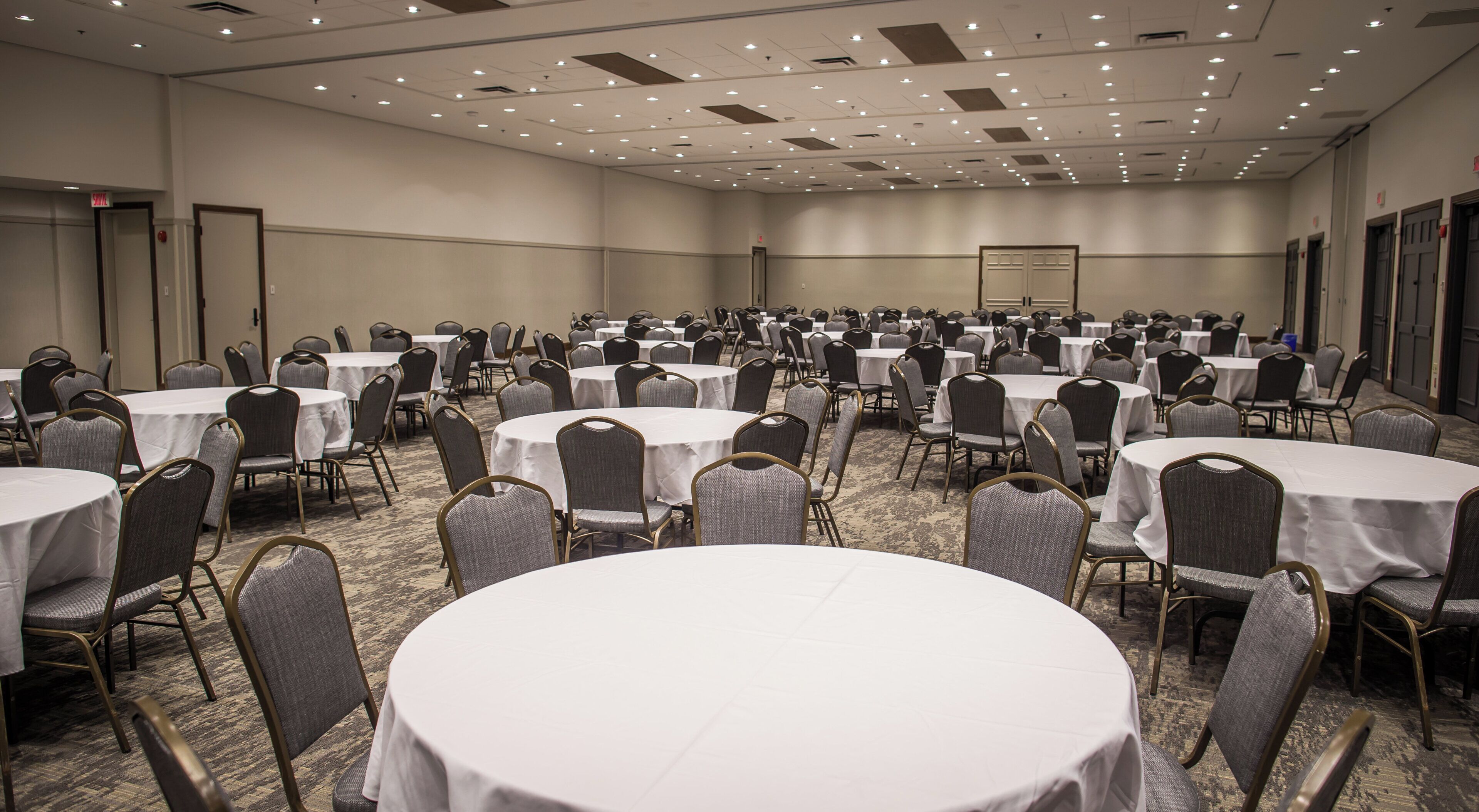 A large conference room with multiple round tables and chairs, carpeted flooring, and a high ceiling with recessed lighting.