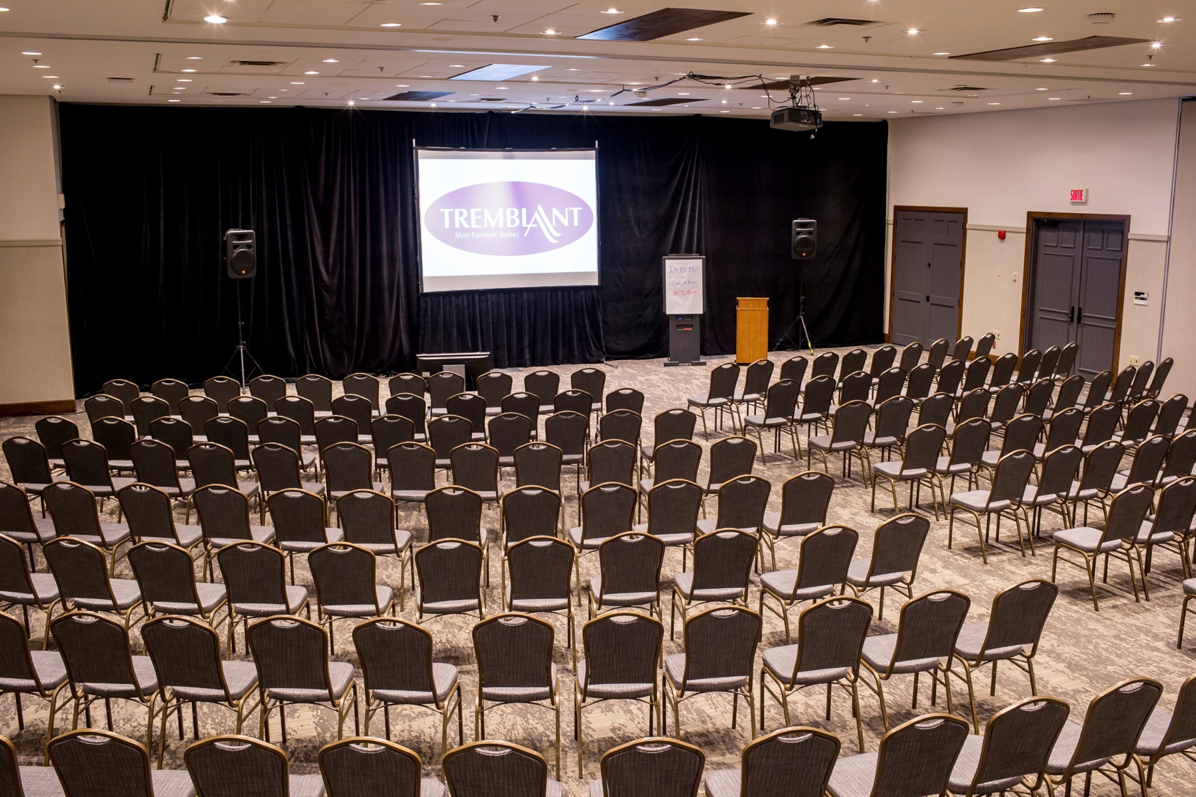 Une salle de conférence avec des rangées de chaises, une scène sur laquelle est installé un écran affichant la mention « TREMBLANT, Mont-Tremblant, Québec », et un pupitre.