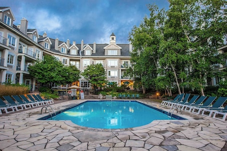 A swimming pool with lounge chairs, surrounded by stone tiles, and a building with multiple balconies in the background.