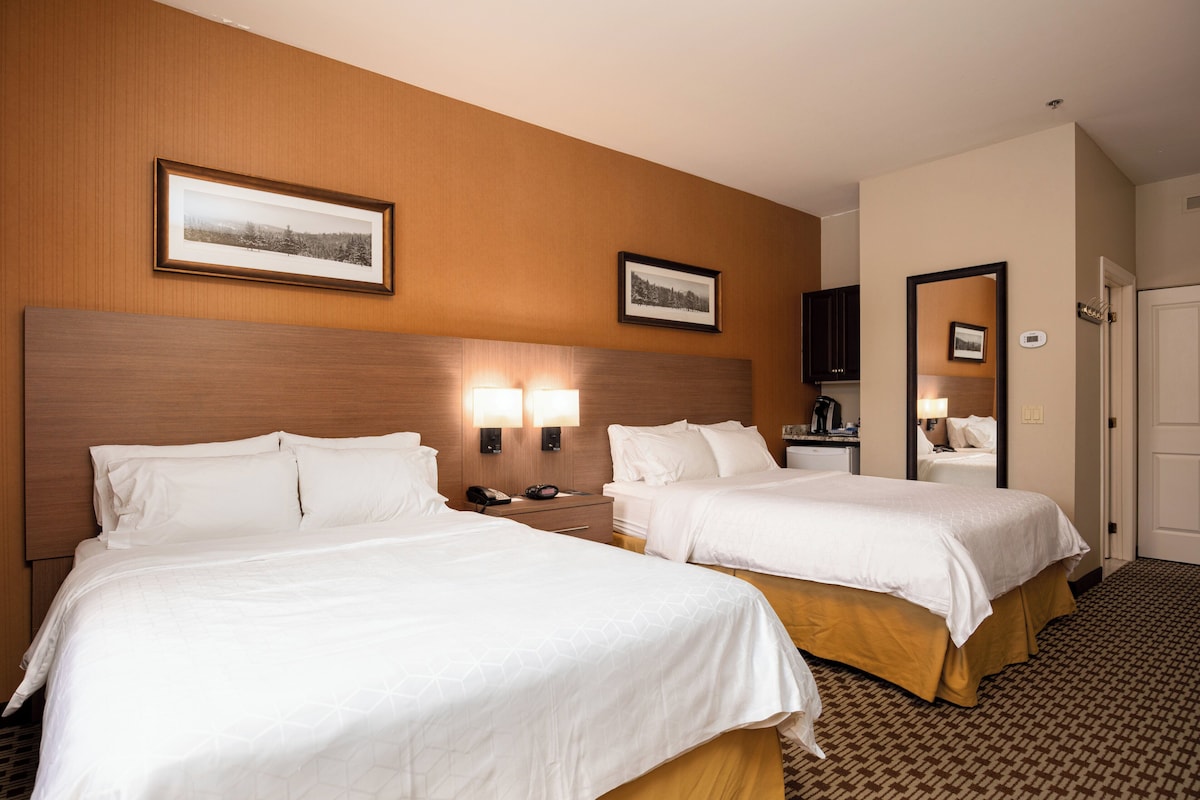 Une chambre d’hôtel avec deux lits, une tête de lit en bois, des lampes fixées au mur et des tableaux encadrés accrochés au mur.
