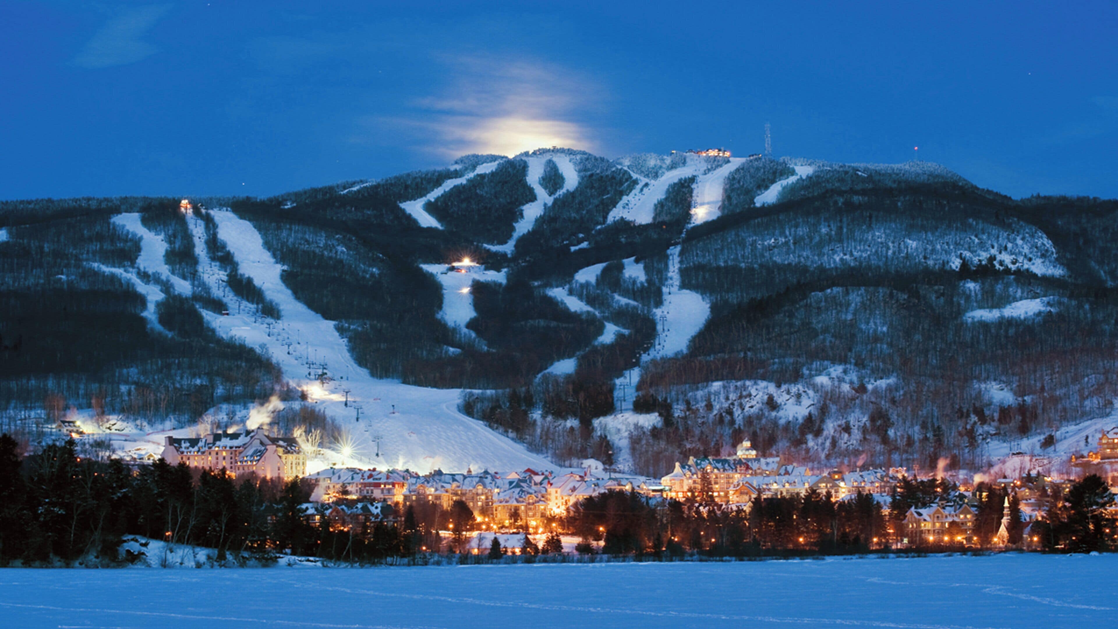 Une montagne enneigée, avec une ville à ses pieds, où l’on peut admirer des bâtiments illuminés et un télésiège.