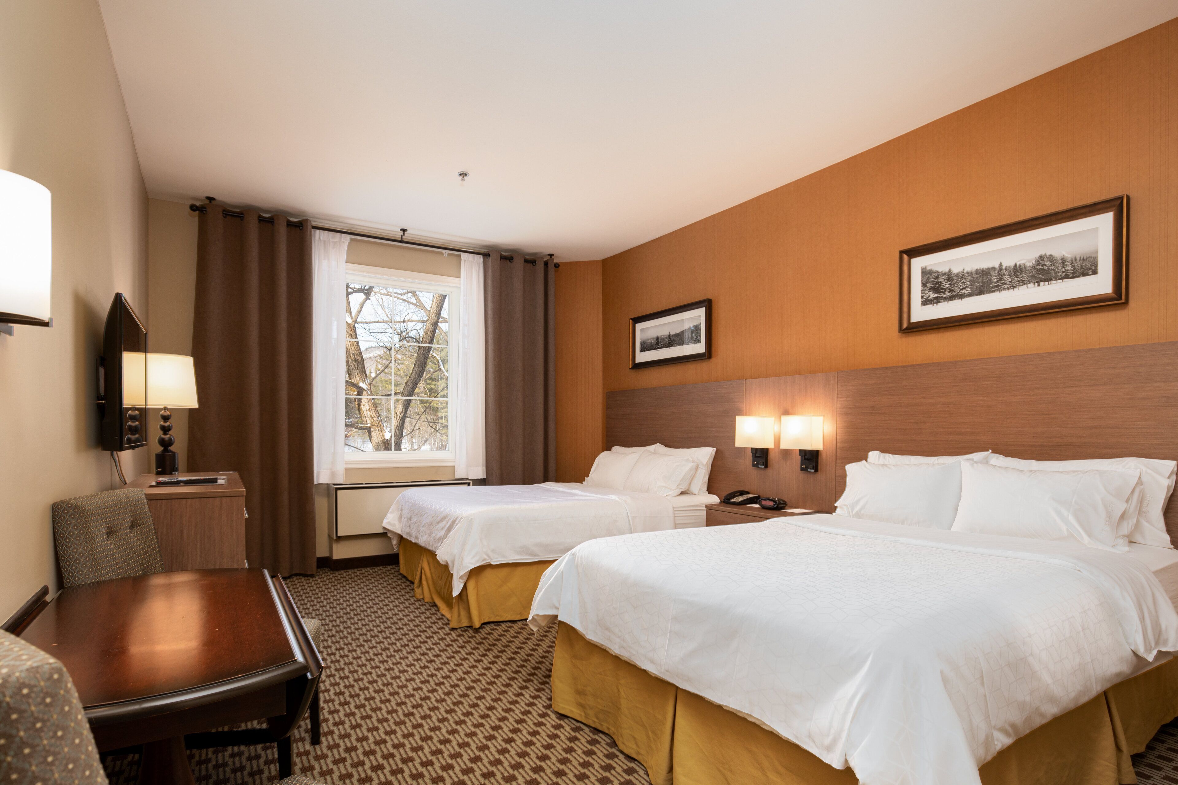 A hotel room with two beds, a desk, a chair, a lamp, and a window with curtains.