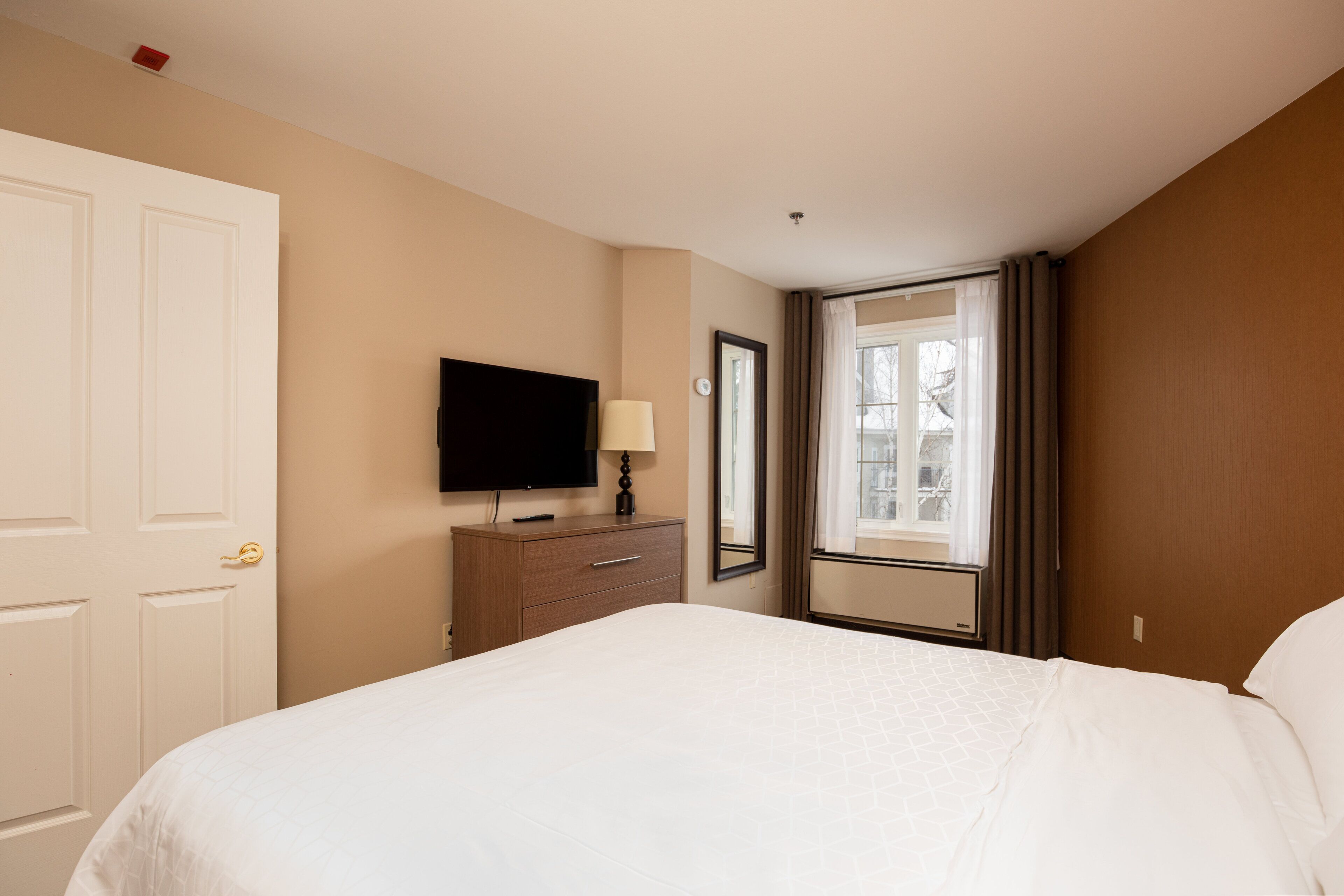 A hotel room with a bed, a television on a dresser, a window with curtains, and a door.