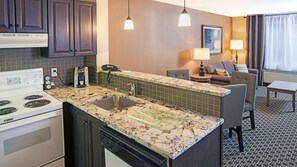 A compact kitchen with a stove, sink, and coffee maker, adjacent to a living area with a sofa and lamp.