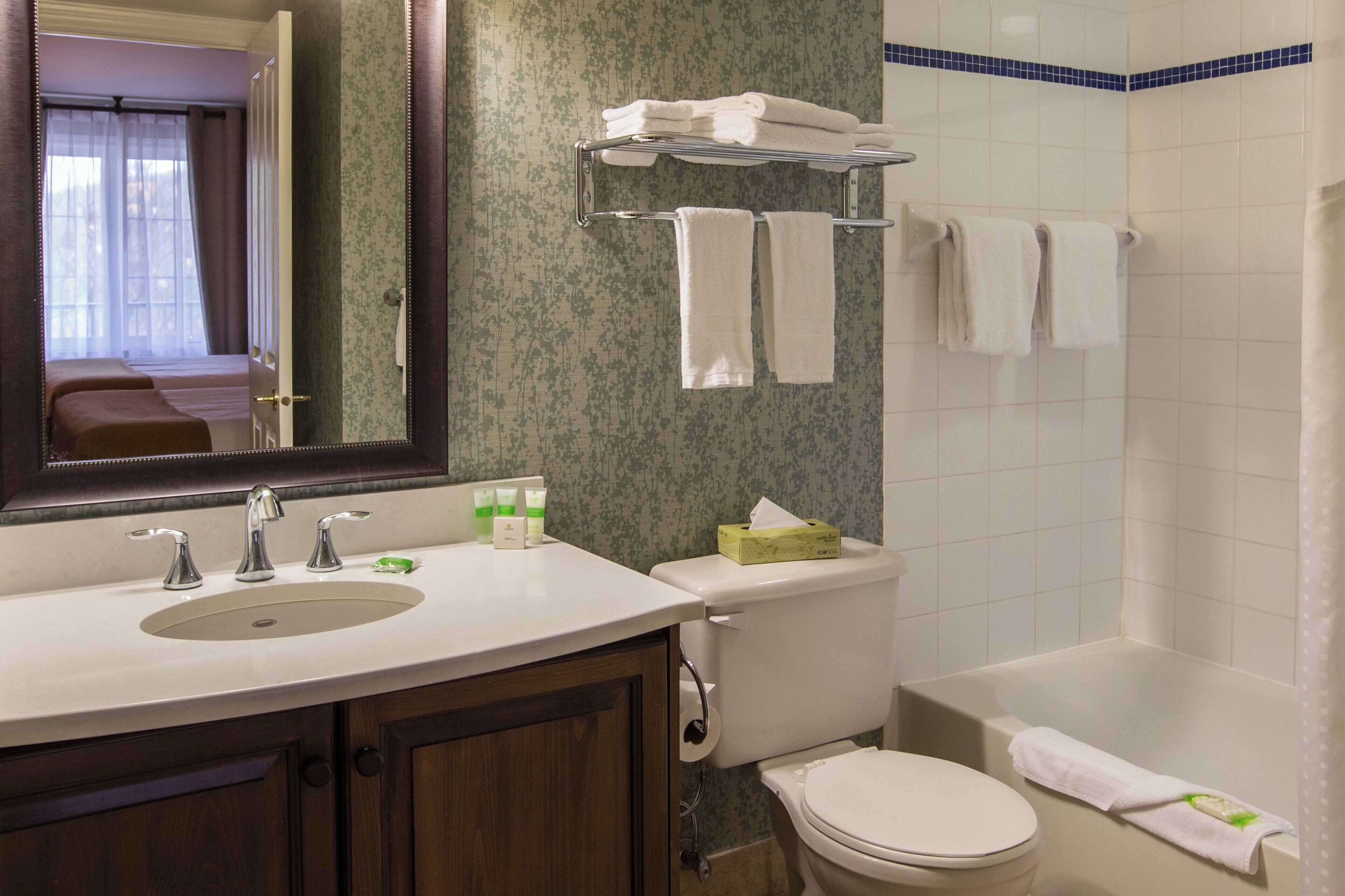 Un baño con lavamanos, inodoro y bañera. Hay toallas en un tendedero y un espejo sobre el lavamanos.