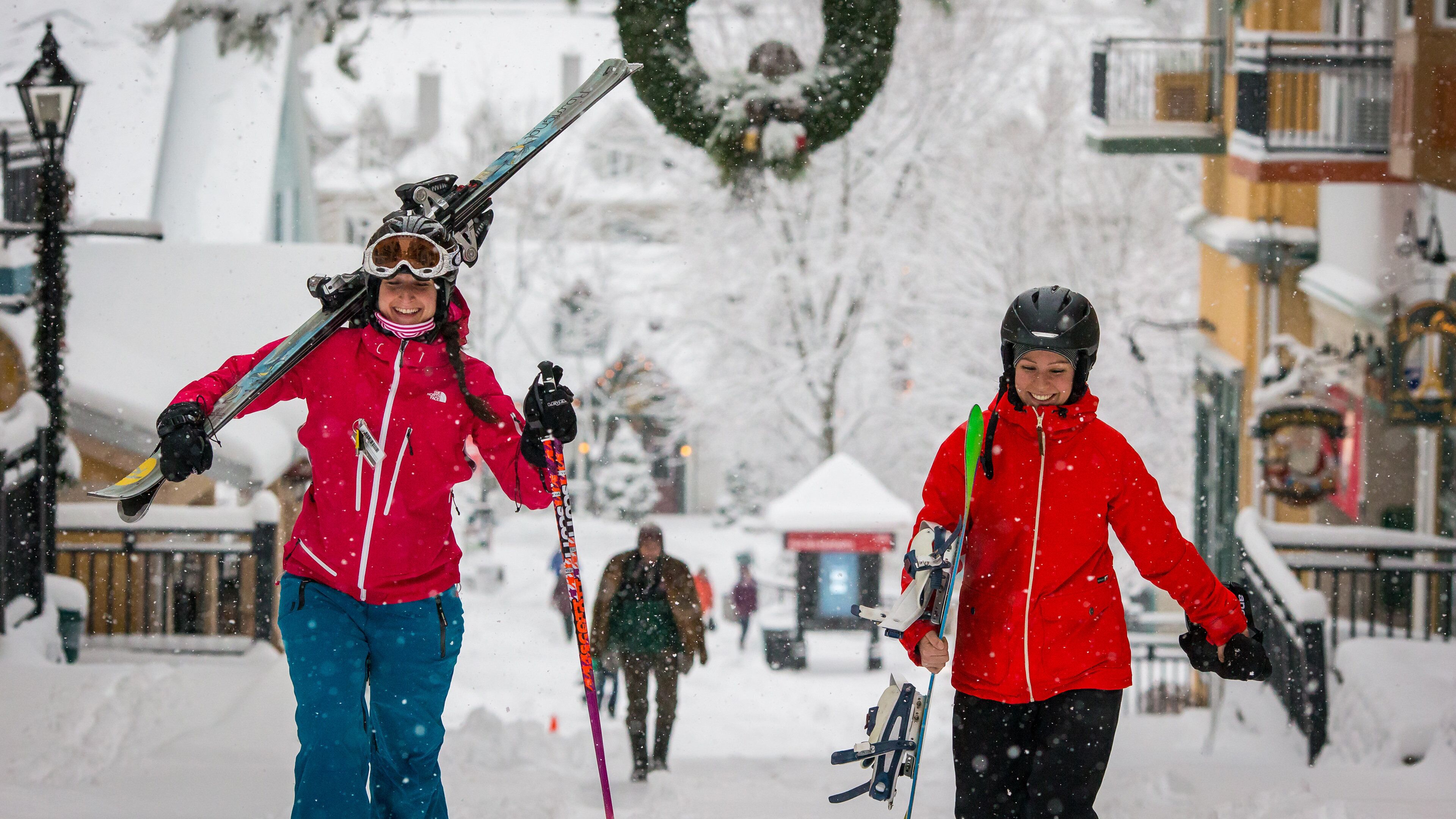 Two individuals with skis, one wearing a red jacket and blue pants, and the other in a red jacket and black pants, walking on a snow-covered street.