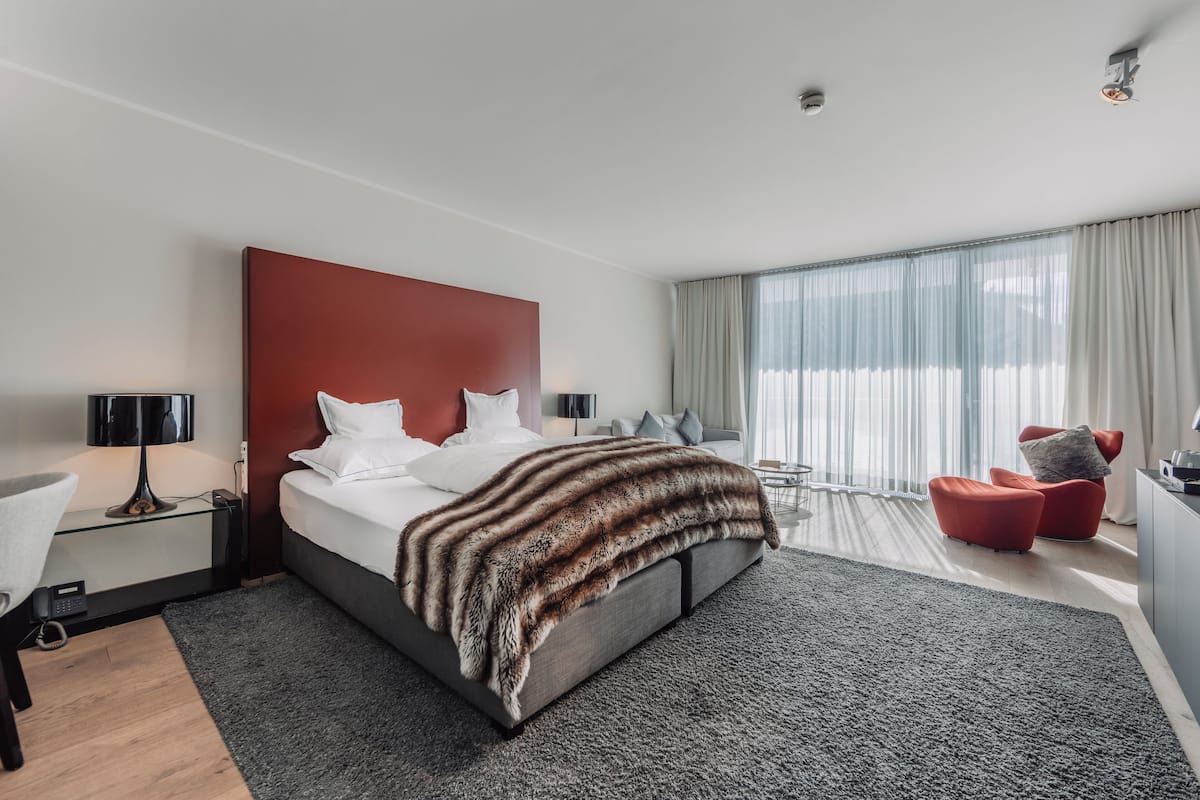 A modern hotel room with a large bed, a desk, a chair, and a large window with curtains.