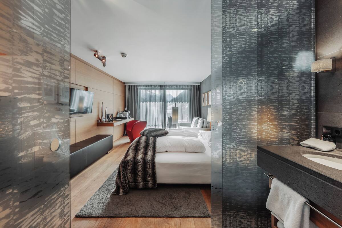 A modern hotel room with a bed, a TV, a desk, and a bathroom with a sink and mirror.