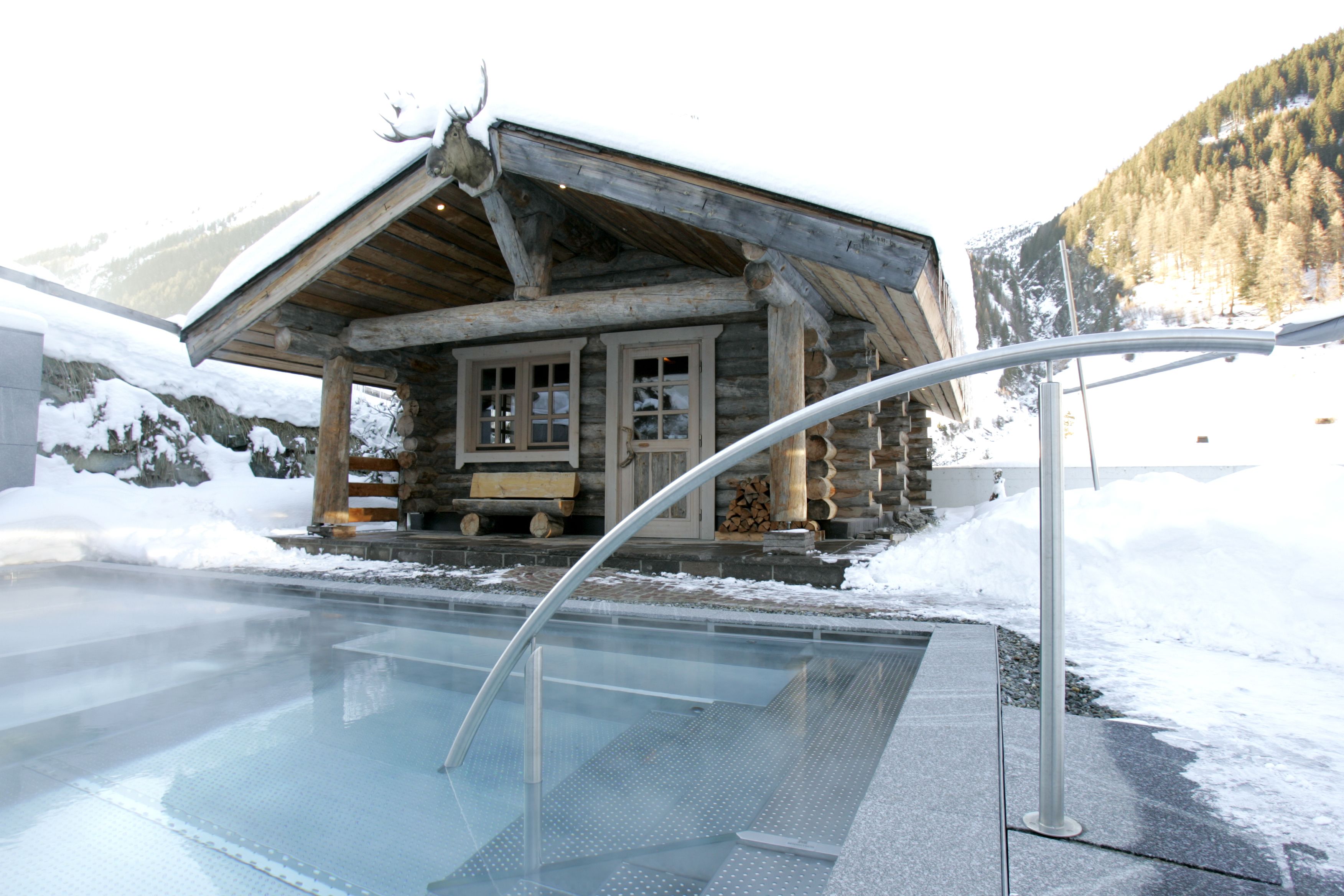 A log cabin with a glass-enclosed hot tub area, surrounded by snow and mountains.