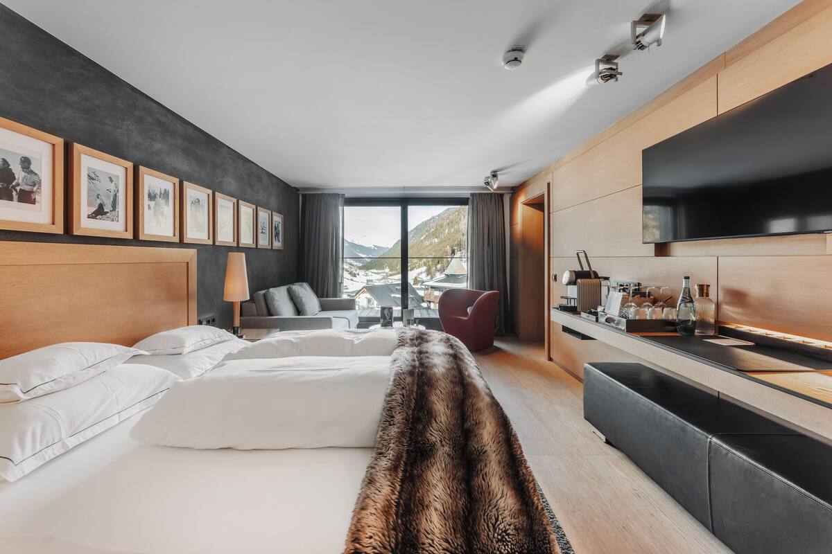 A modern hotel room with a large bed, a sofa, a dining area, and a view of mountains.