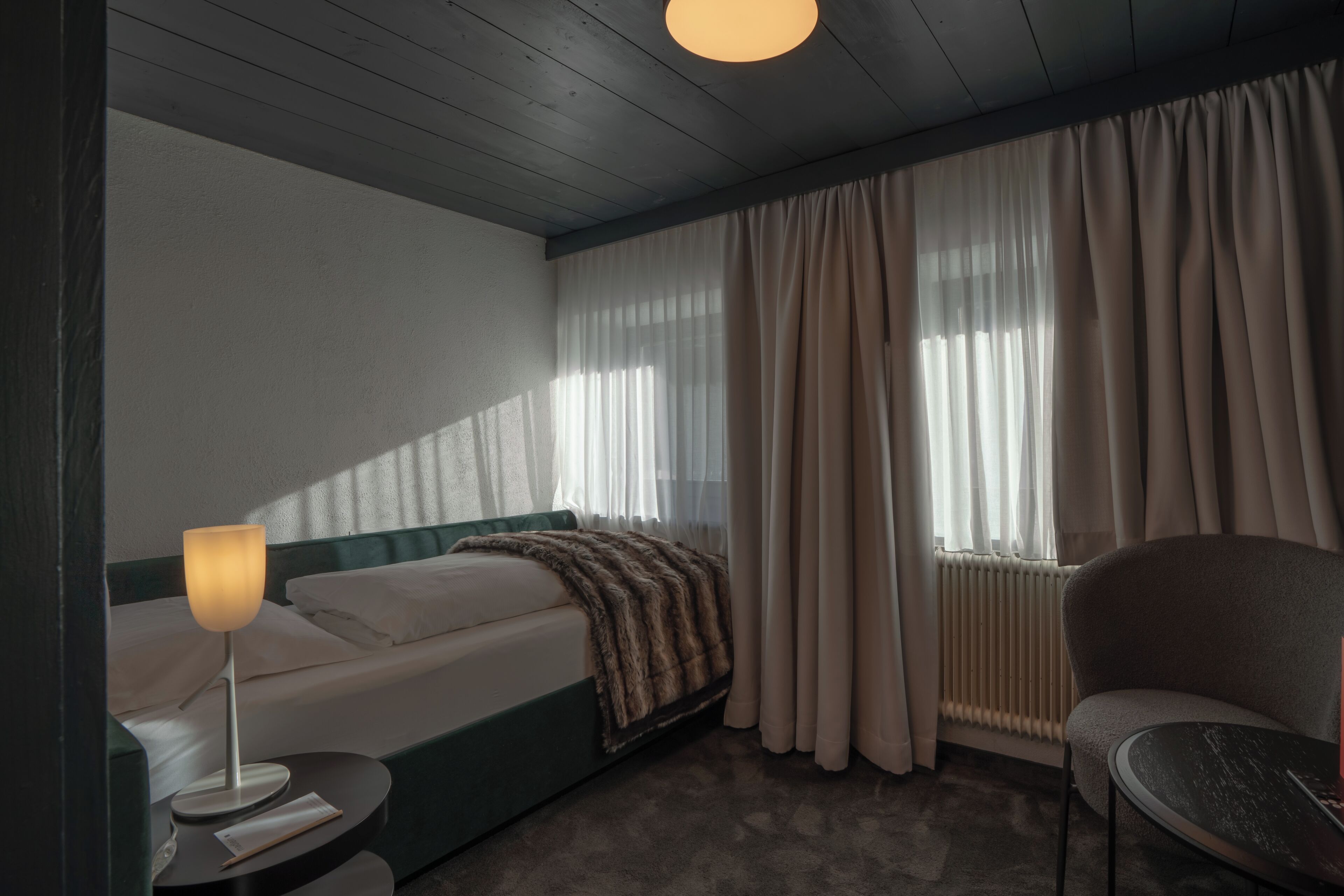 Una habitación de hotel con cama, mesita de noche con lámpara, una silla y ventana con cortinas.