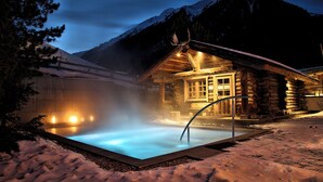Una cabaña de troncos con un jacuzzi rodeada de nieve, con montañas al fondo.