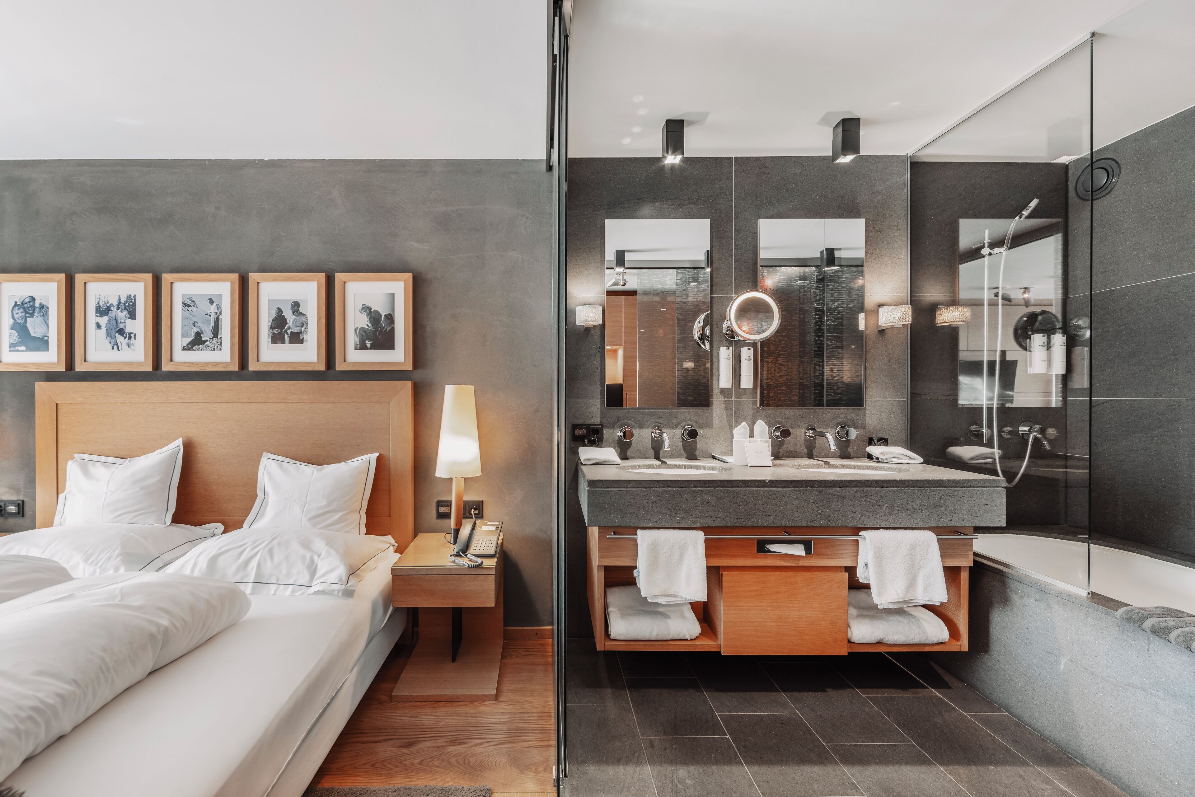 A modern hotel room with a bed, bedside table, lamp, framed pictures, and a bathroom with a large mirror, multiple sinks, and a bathtub.