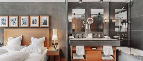 A modern hotel room with a bed, bedside table, lamp, framed pictures, and a bathroom with a large mirror, multiple sinks, and a bathtub.