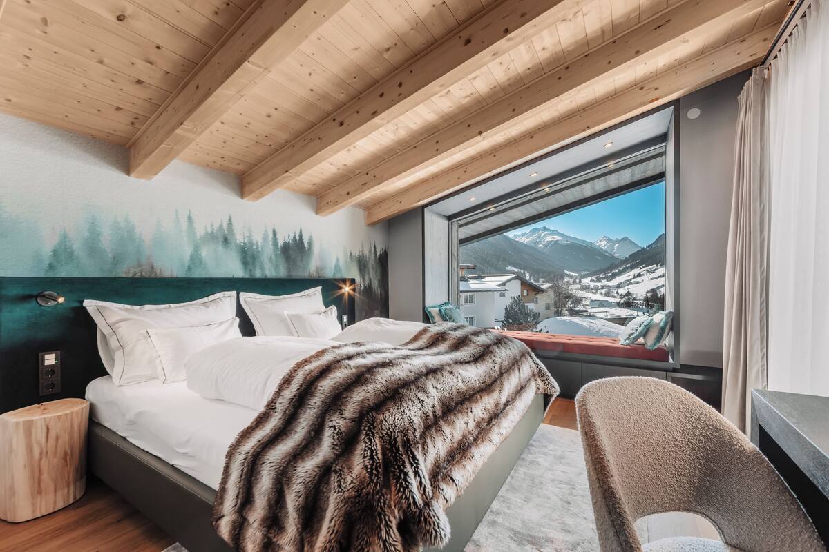 A bedroom with a bed, a view of mountains, and a wooden ceiling.