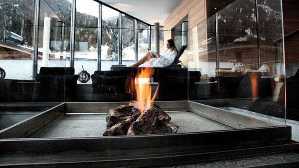 Un interior moderno con chimenea de vidrio, una persona relajándose junto al fuego y vistas a un paisaje montañoso cubierto de nieve.