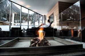 A modern interior with a glass fireplace, a person relaxing by the fire, and a view of a snowy mountain landscape. - Hotel Madlein (Ischgl)