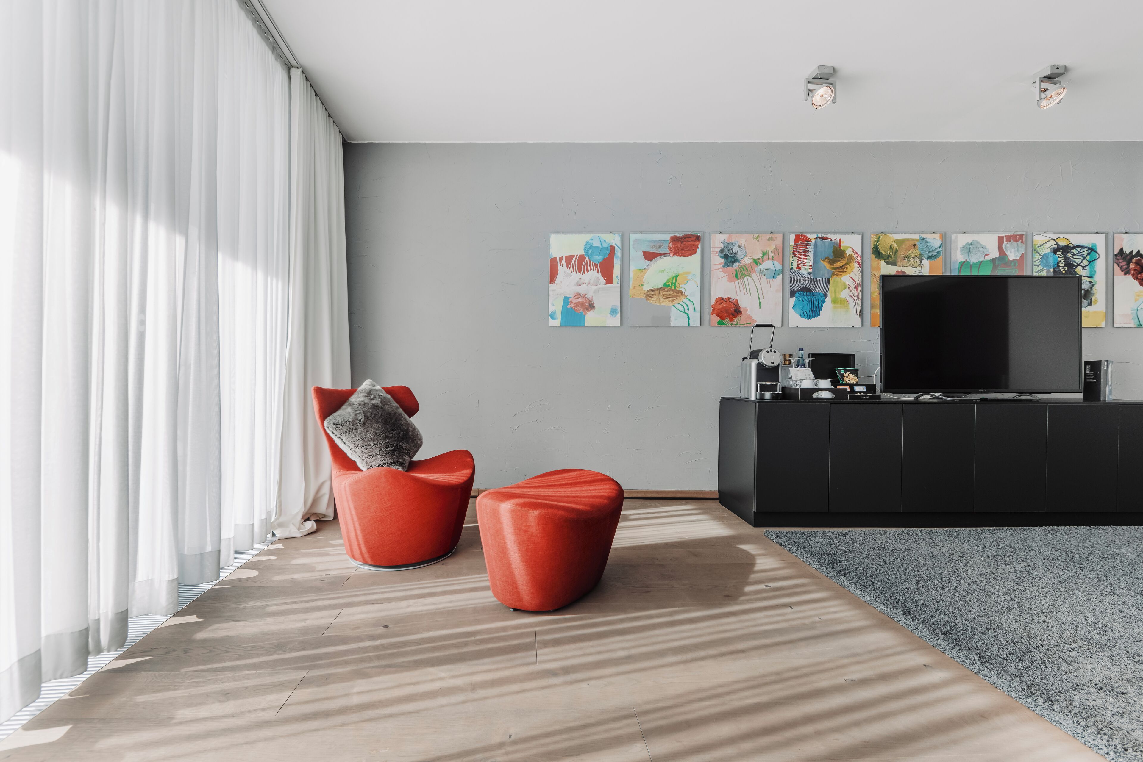Un salon moderne doté d’un grand téléviseur à écran plat, de fauteuils rouges, d’œuvres d’art abstraites accrochées au mur et d’un tapis gris.