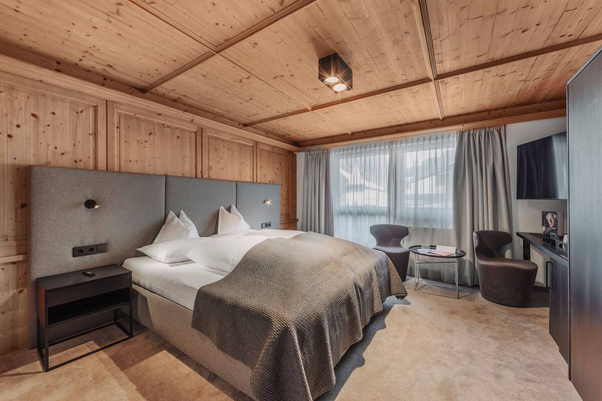 A hotel room with a wooden ceiling, a large bed with white bedding, a small bedside table, a chair, a desk, and a television.