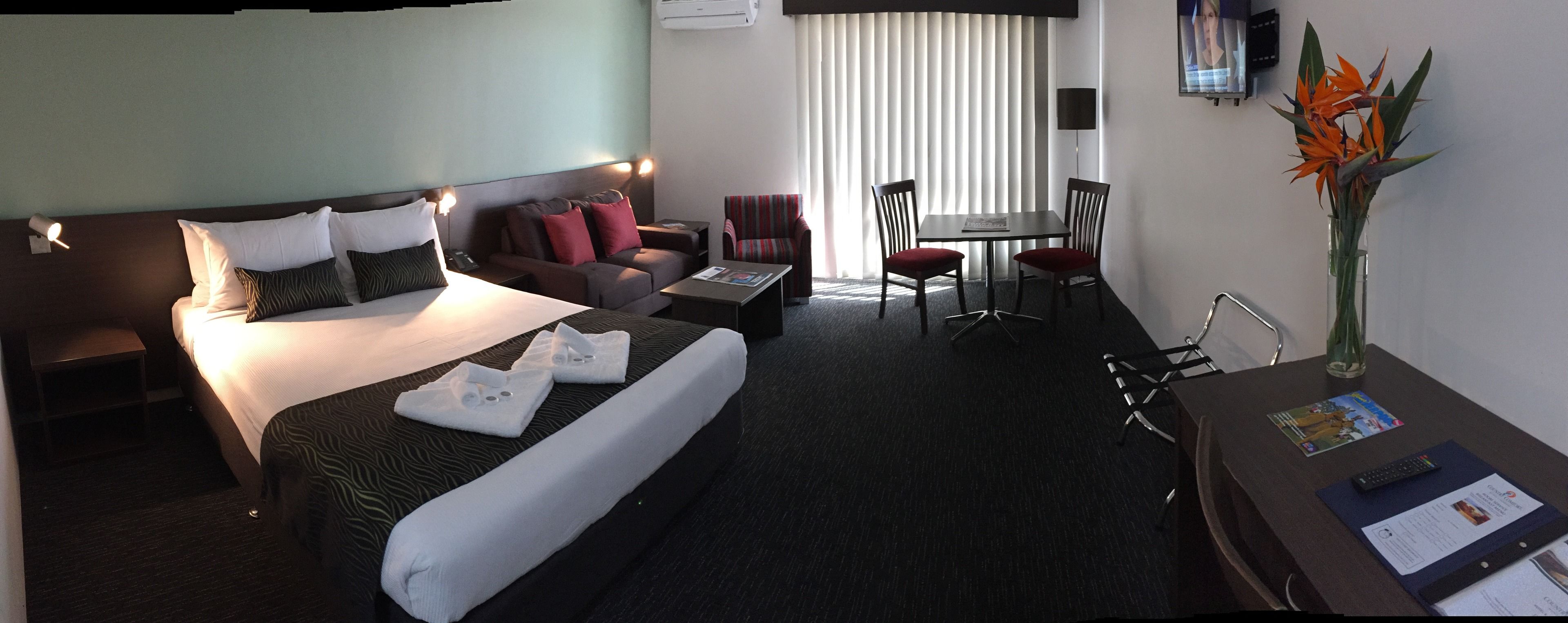 A hotel room with a bed, a sofa, a desk, and a chair. There is a TV on the wall and a window with blinds.
