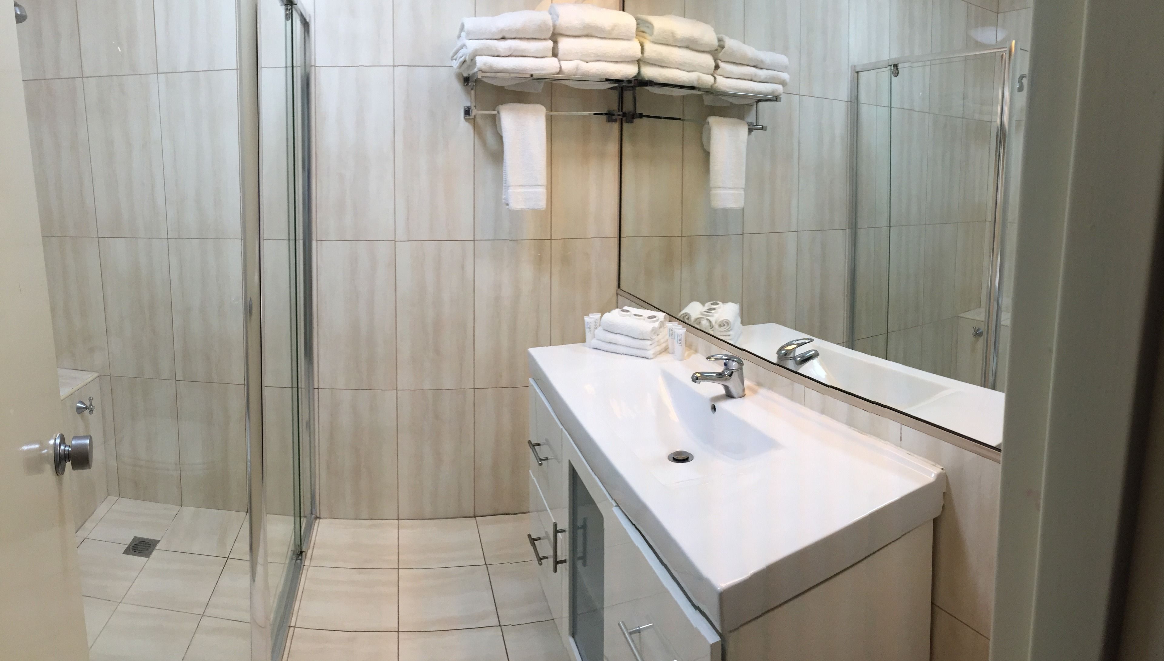 Un baño moderno con cabina de ducha de vidrio, un lavabo blanco con espejo y toallas dobladas ordenadamente en un estante.