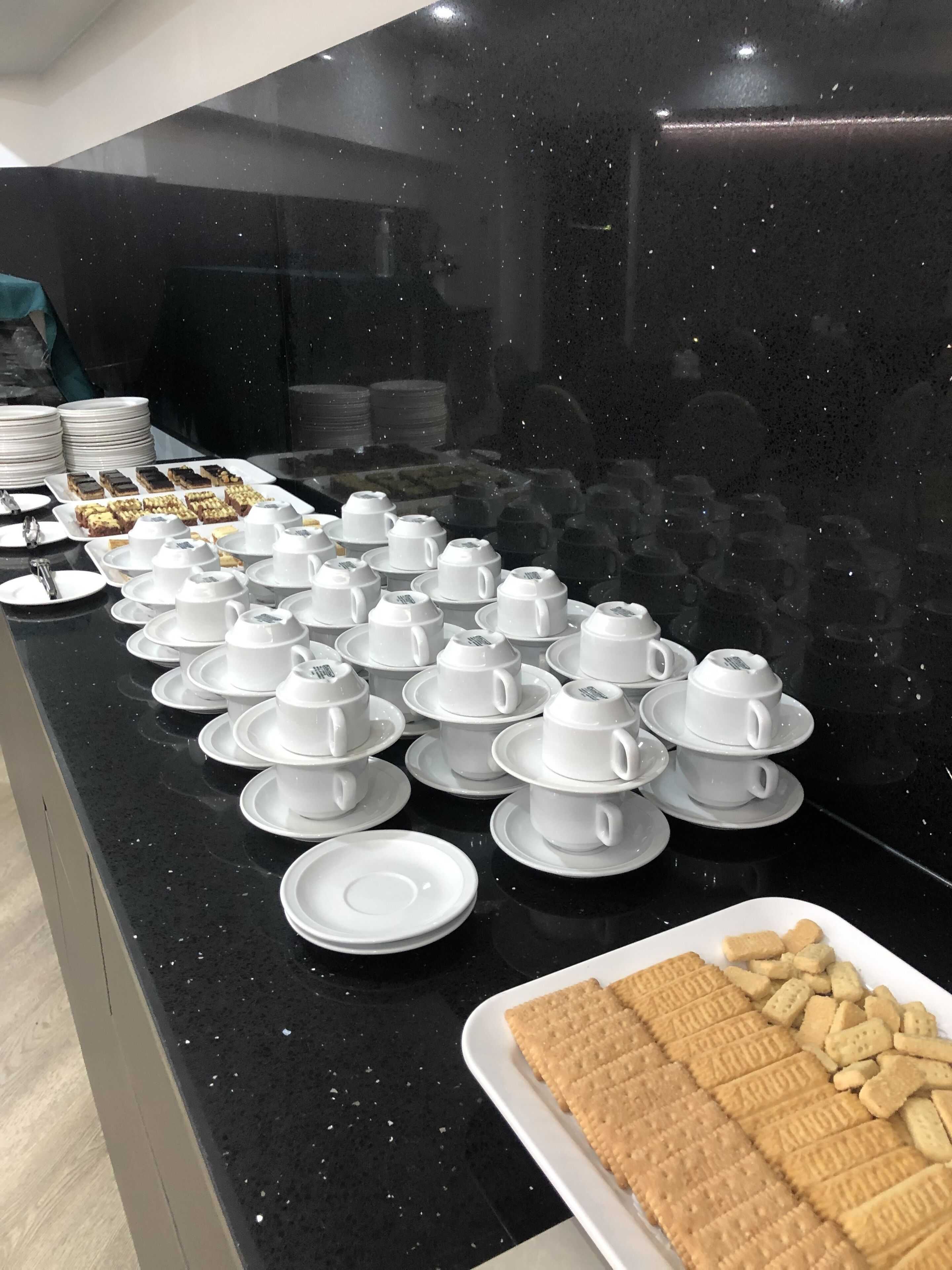 Una barra de desayuno con varias tazas y platillos blancos, un plato de galletas y pilas de platos.