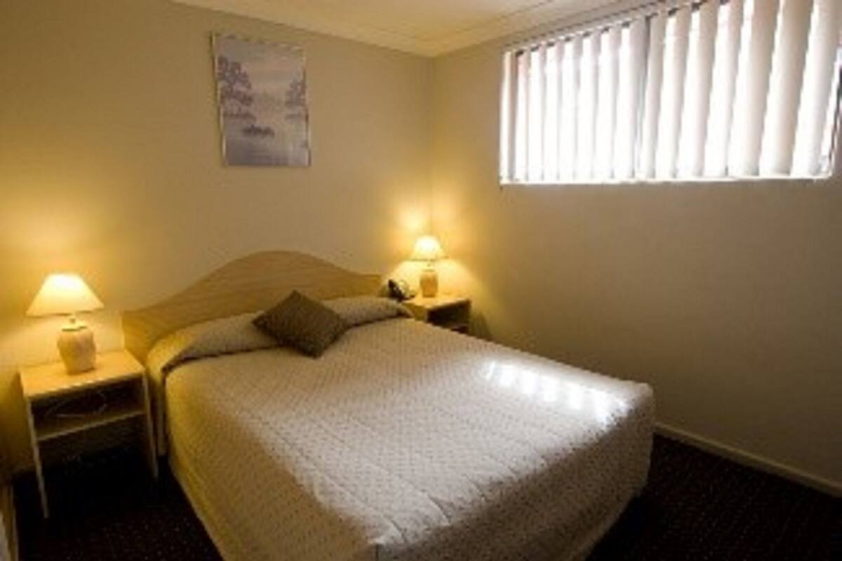 Ein Hotelzimmer mit einem großen Bett, zwei Nachttischen mit Lampen, einem Fenster mit Jalousien und einem Wandbild.