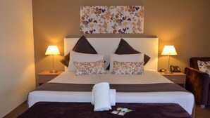 A neatly made bed with floral pillows, a headboard with a floral artwork, two bedside lamps, and a purple ottoman with a towel and a small item on it. - Hunts Hotel Liverpool (Casula)