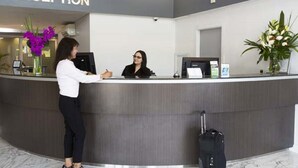 A reception desk with a receptionist, a guest standing, and a black suitcase on the floor. - Hunts Hotel Liverpool (Casula)