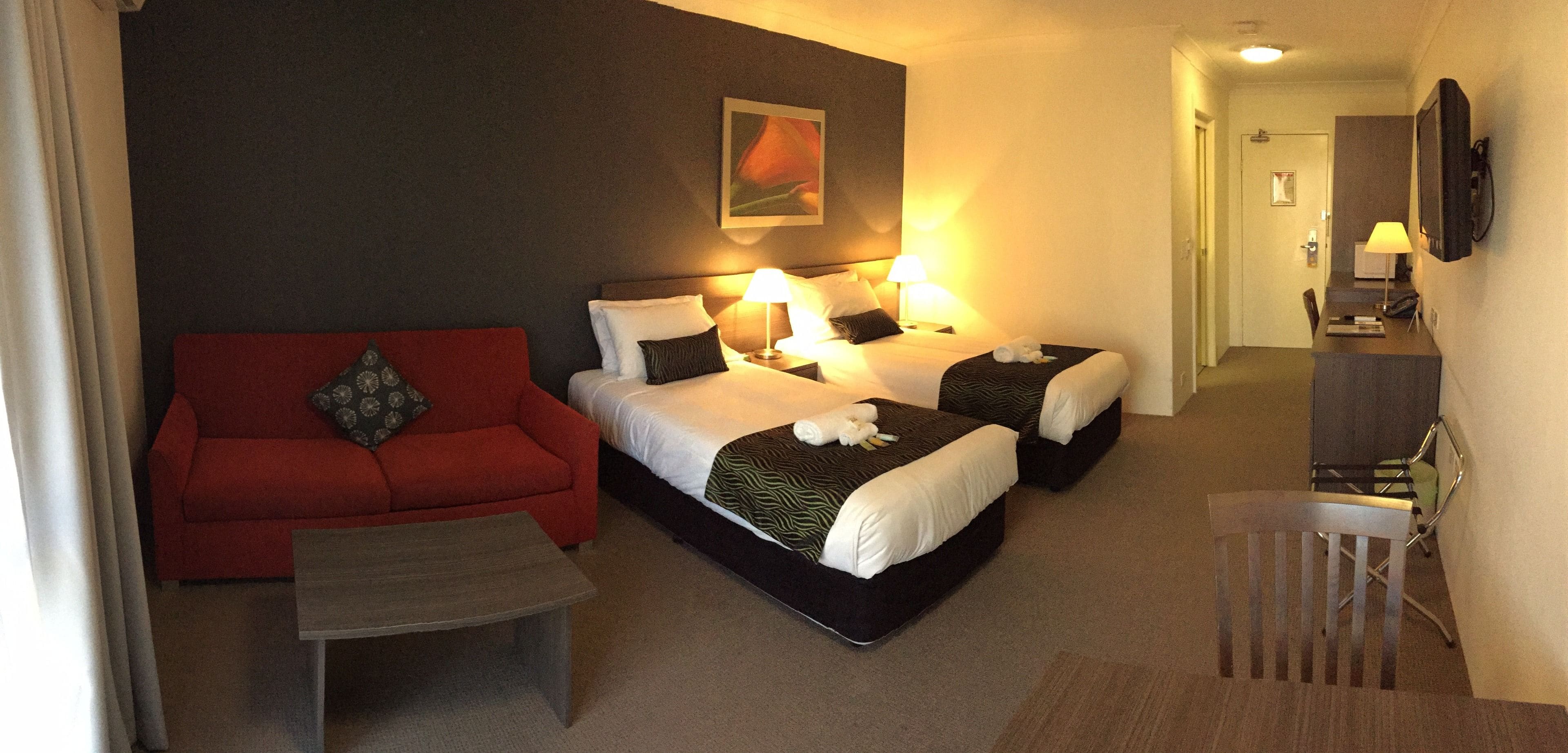 Een hotelkamer met een rode bank, twee bedden met witte lakens, een kleine tafel en een bureau met een lamp.