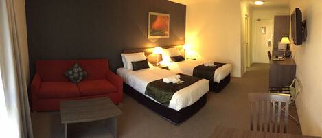 Een hotelkamer met een rode bank, twee bedden met witte lakens, een kleine tafel en een bureau met een lamp.