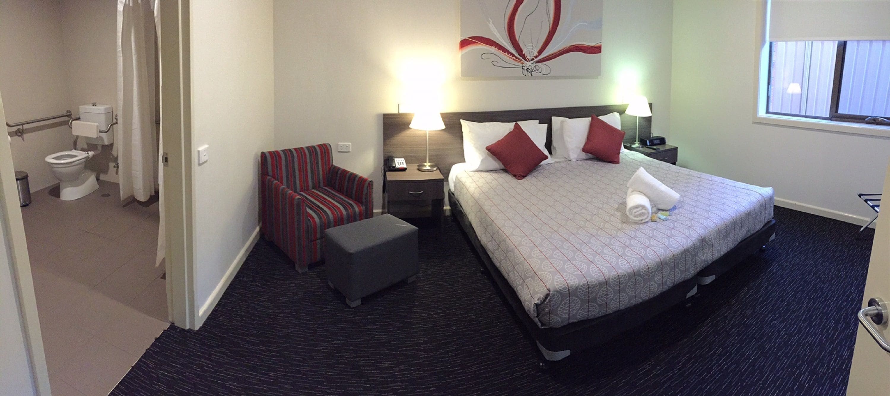 A hotel room with a bed, a chair, a nightstand, a lamp, and a bathroom with a toilet and shower.