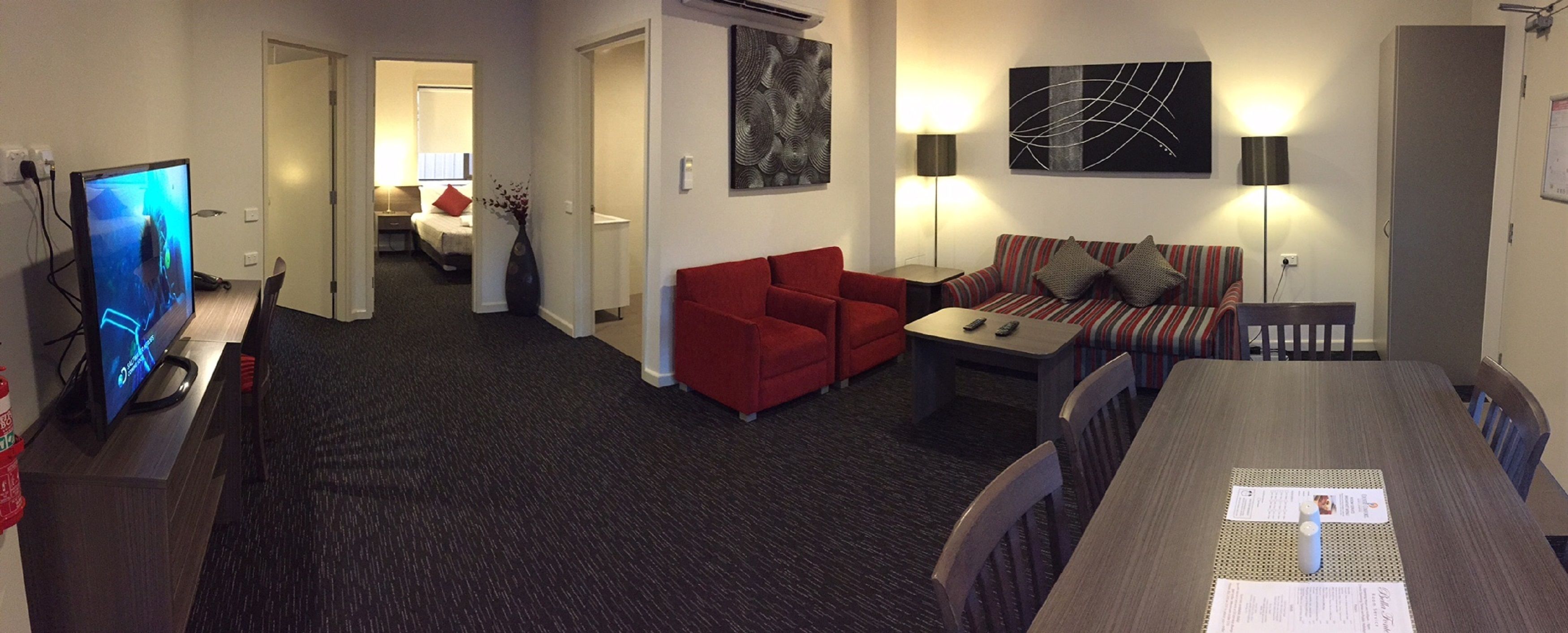 Une chambre d’hôtel équipée d’une télévision, d’un canapé rouge, d’un canapé à rayures, d’une table à manger et d’œuvres d’art accrochées aux murs.