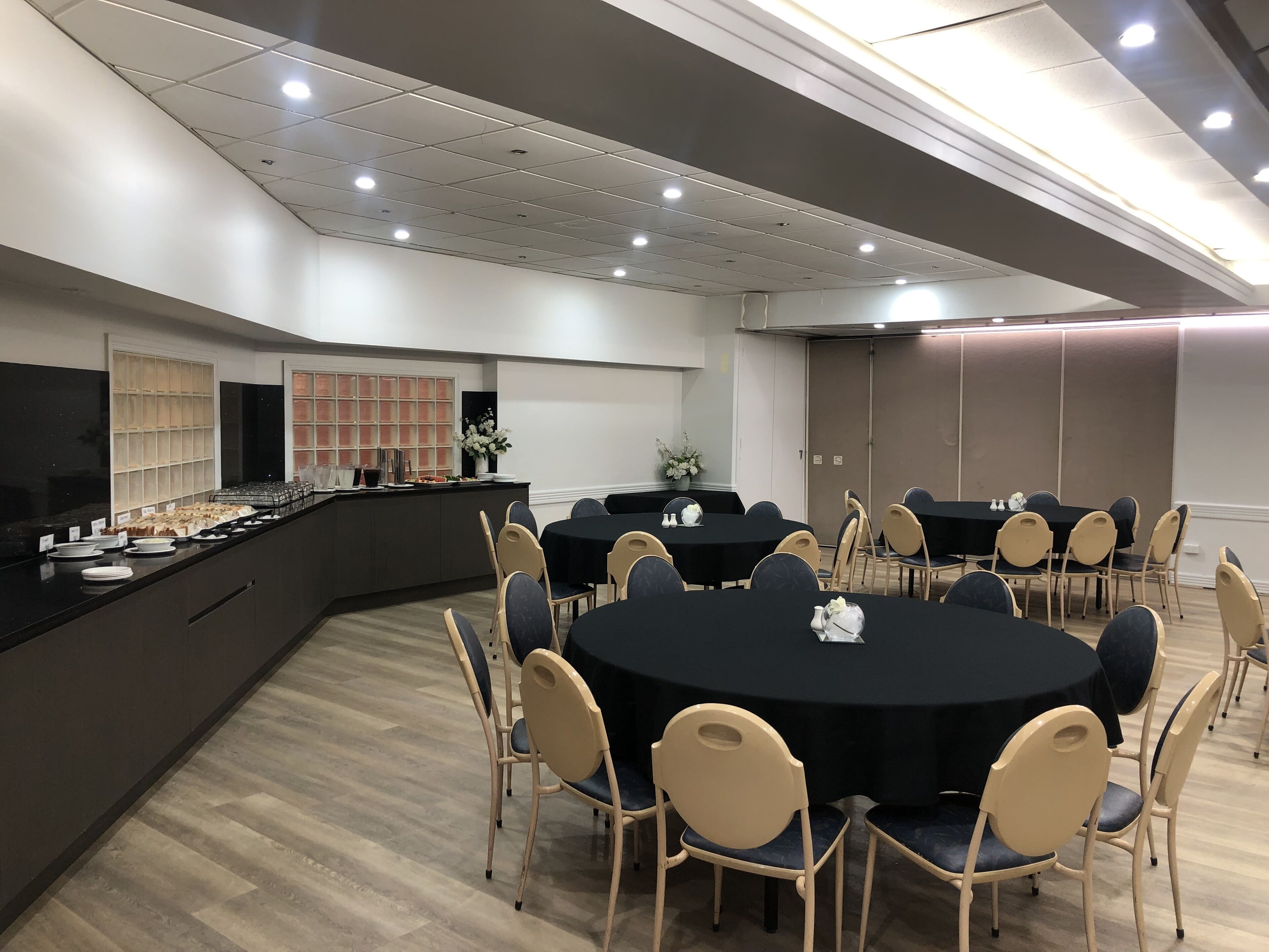 Une salle de conférence équipée de tables rondes, de chaises de couleur beige, d’un espace buffet et d’un grand tableau blanc.