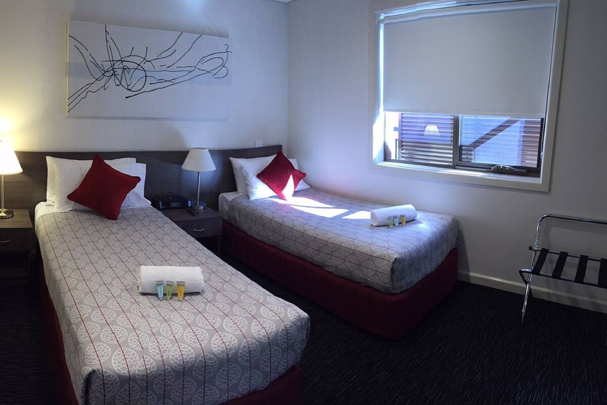 Ein Hotelzimmer mit zwei Einzelbetten, einem Nachttisch mit Lampe, einem Wandbild, einem Fenster mit Jalousien und einem kleinen Tisch mit Toilettenartikeln.
