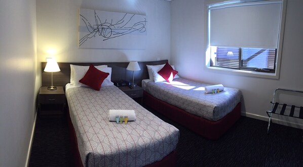 A hotel room with two single beds, a nightstand with a lamp, a wall art piece, a window with blinds, and a small table with toiletries.