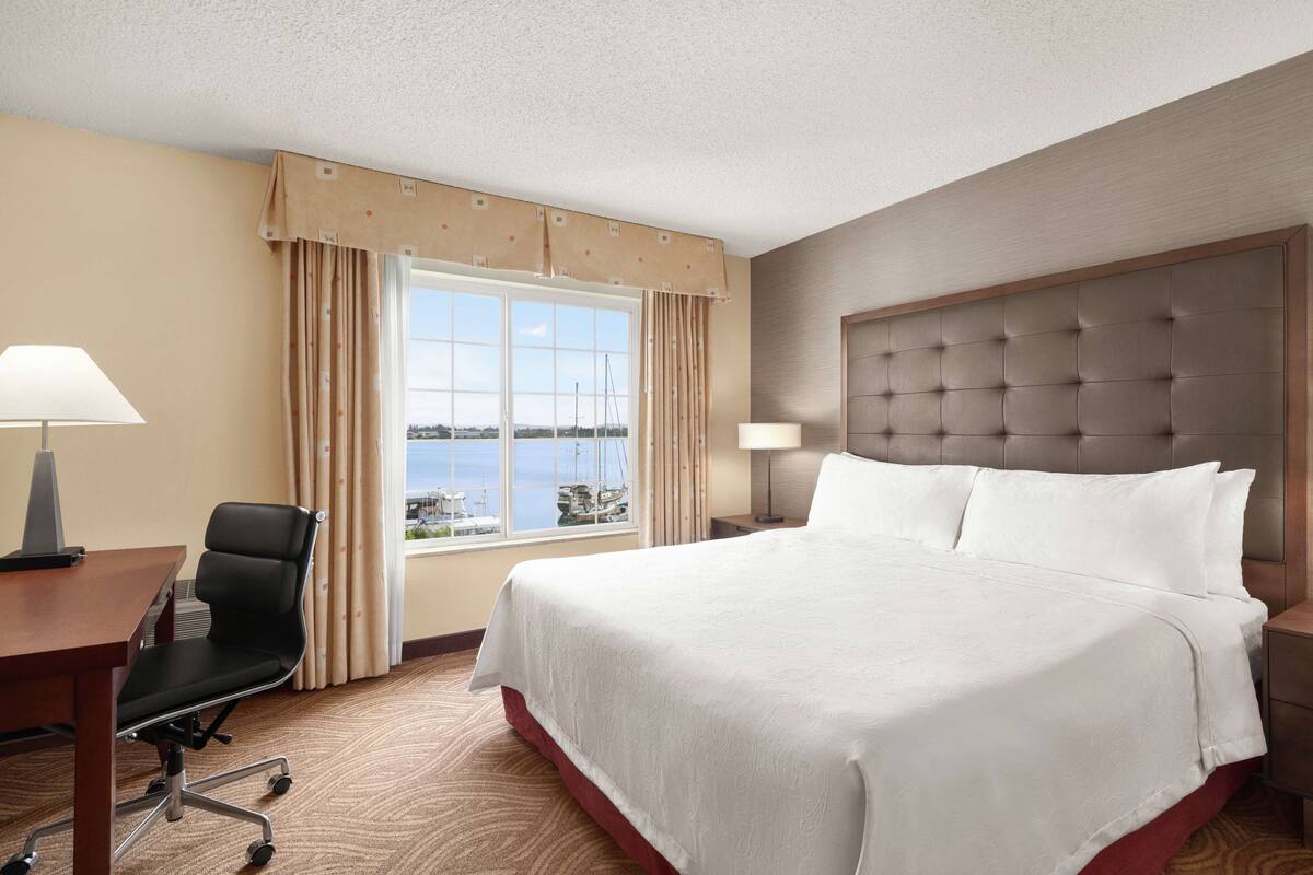 Ein Hotelzimmer mit einem großen Bett, einem Schreibtisch mit Stuhl, einem Fenster mit Vorhängen und Blick aufs Meer.