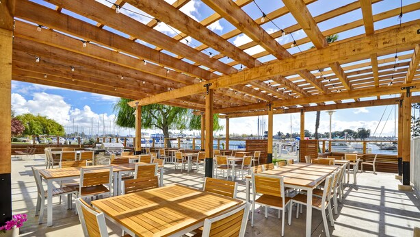 An outdoor patio with wooden tables and chairs, a pergola with string lights, and a view of the marina. - Homewood Suites by Hilton Oakland-Waterfront (Oakland)