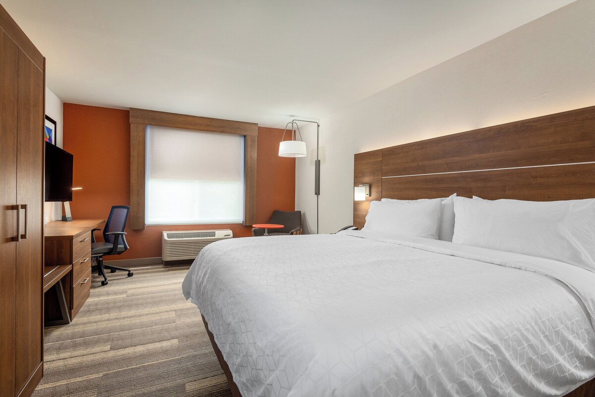 A hotel room with a large bed, a desk with a chair, a TV, and a window with blinds.