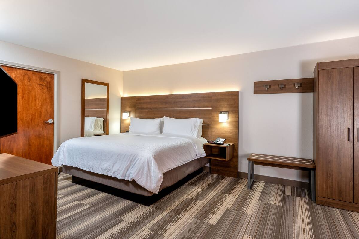 A modern hotel room with a large bed, wooden headboard, bedside tables with lamps, a bench, and a wardrobe.