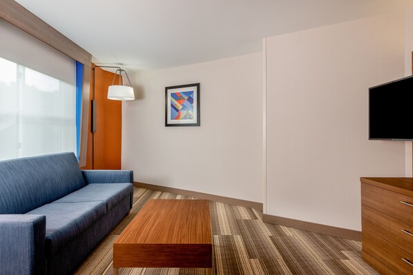 Een moderne hotelkamer met een blauwe bank, een houten salontafel, een lijst met een abstract schilderij en een flatscreen televisie aan de muur.