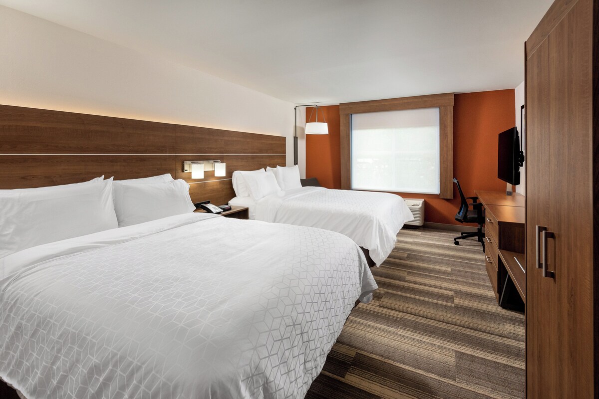 A hotel room with two beds, a desk, a chair, a TV, and a window.