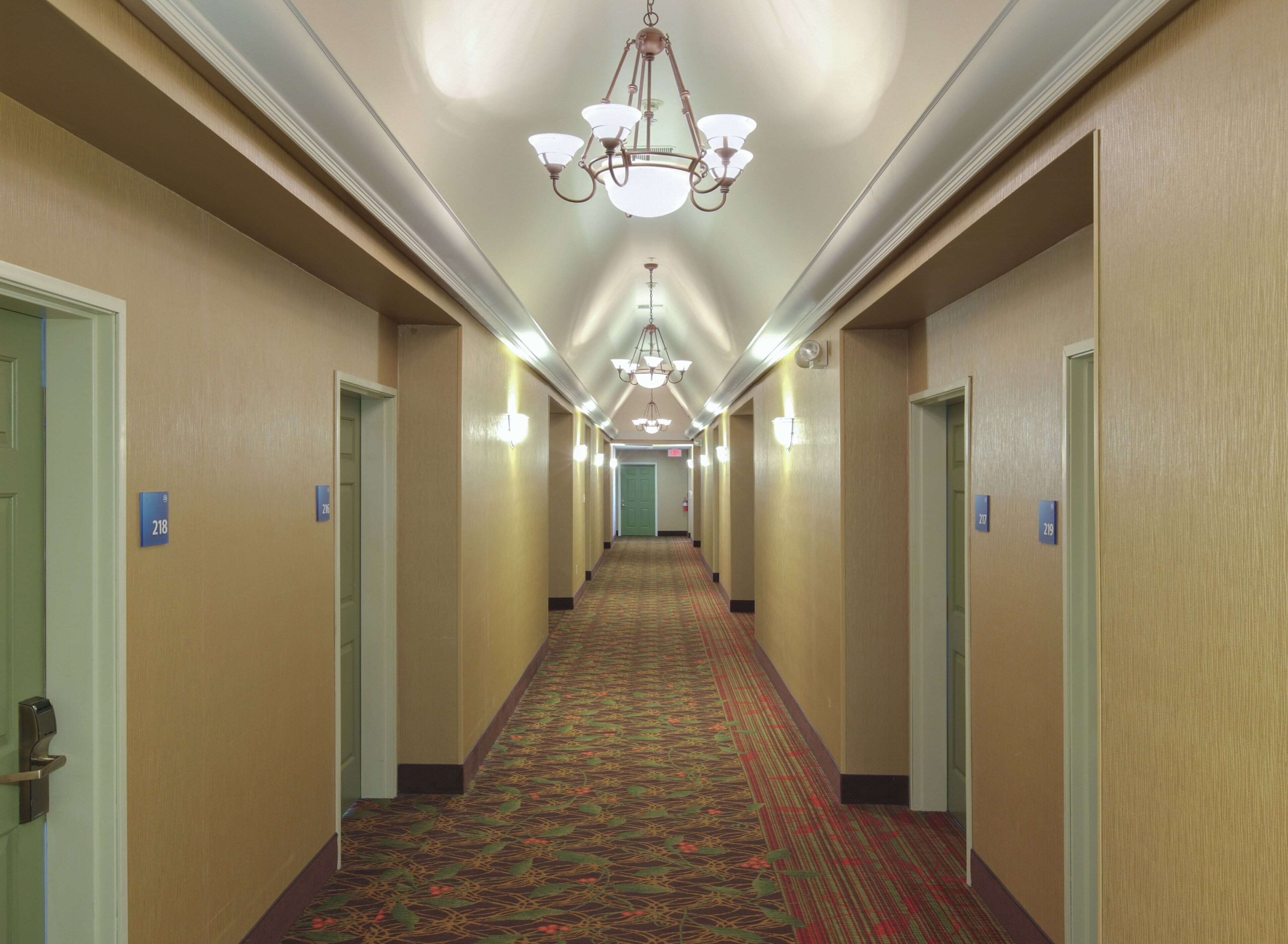 A hotel hallway with carpeted floors, doors with room numbers, and a chandelier.