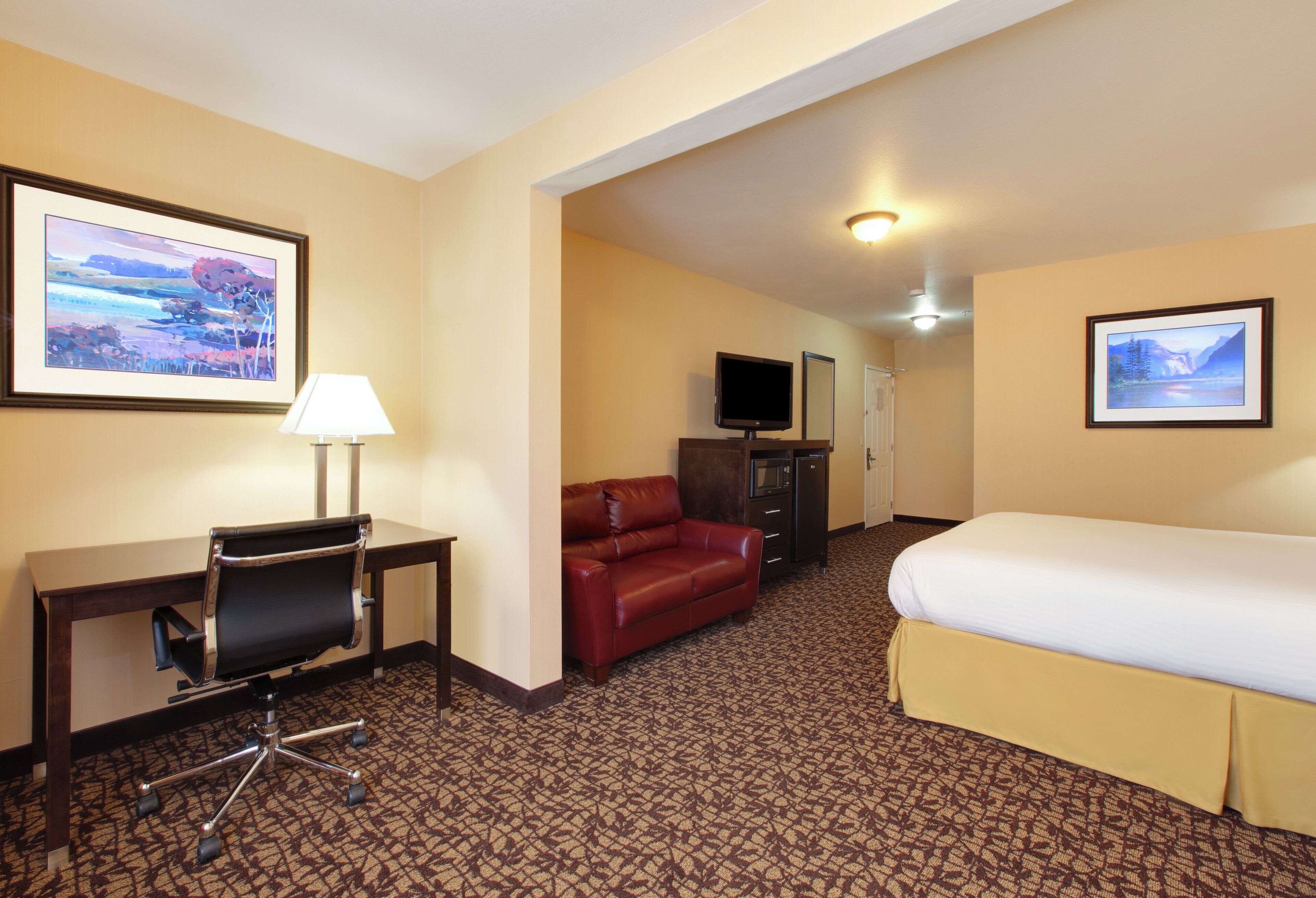 A hotel room with a bed, a desk with a chair, a red sofa, a television, and framed pictures on the walls.